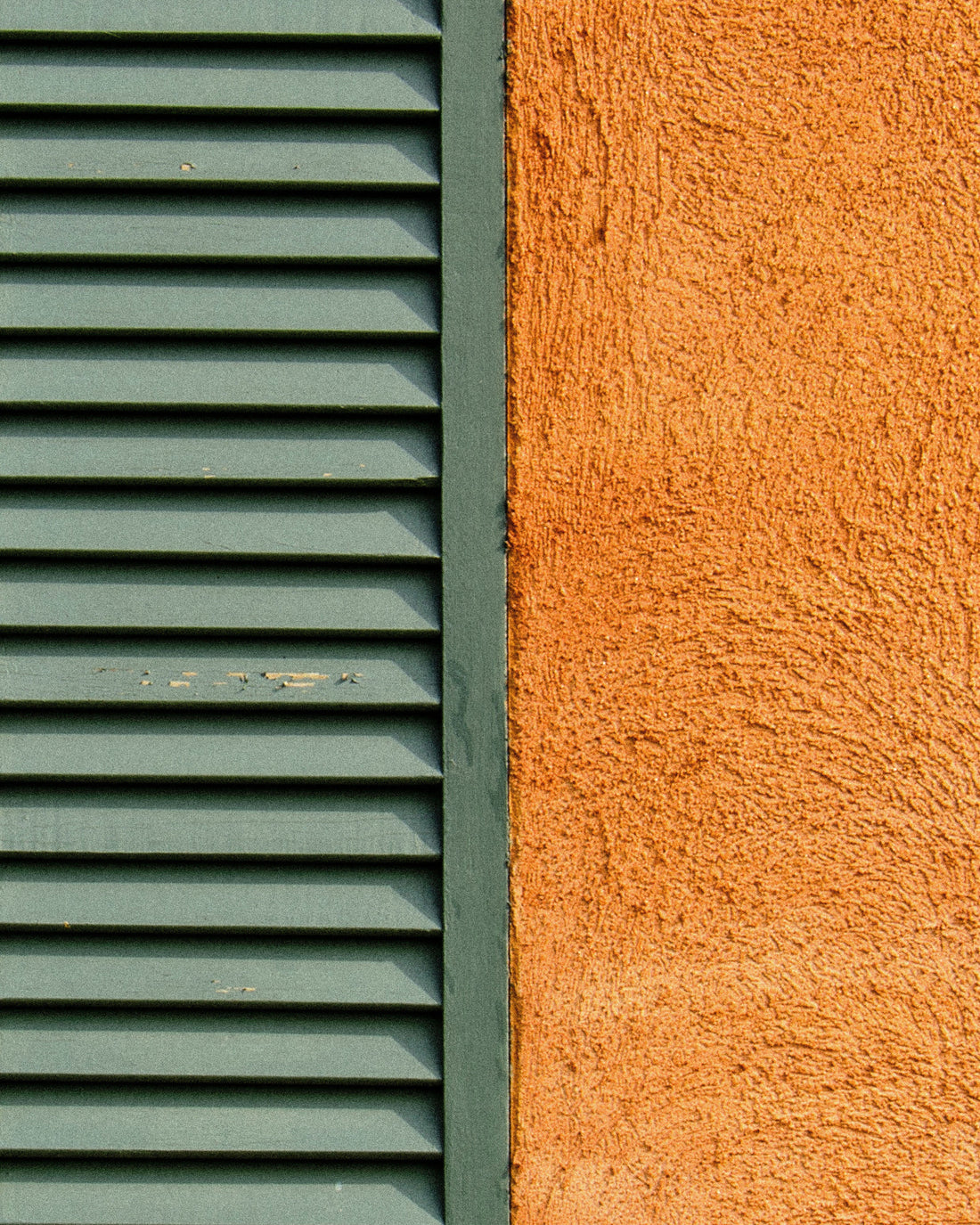 A green louvered wooden shutter beside a rough ochre stucco wall in full sun